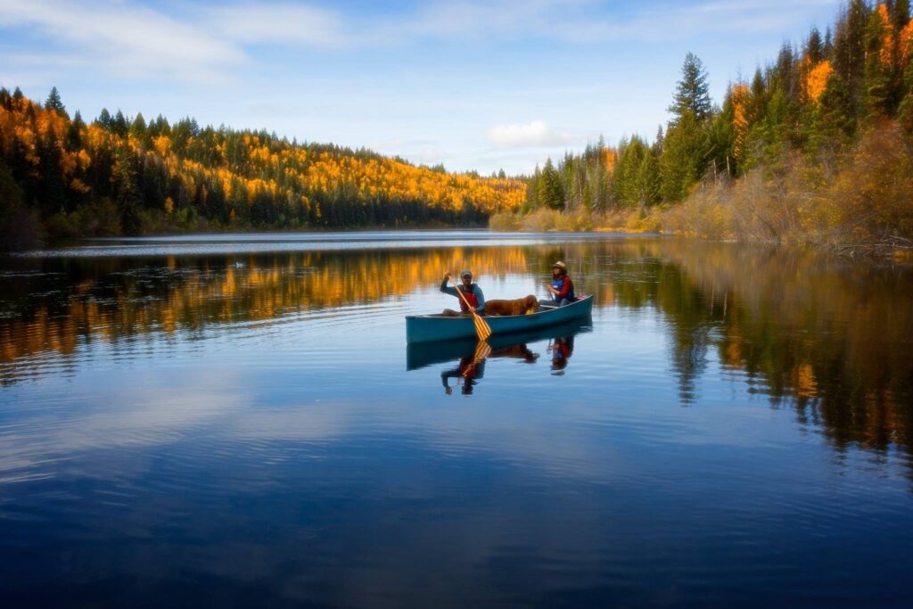 Two paddlers and a dog in a canoe on a calm lake in fall colours near Prince George